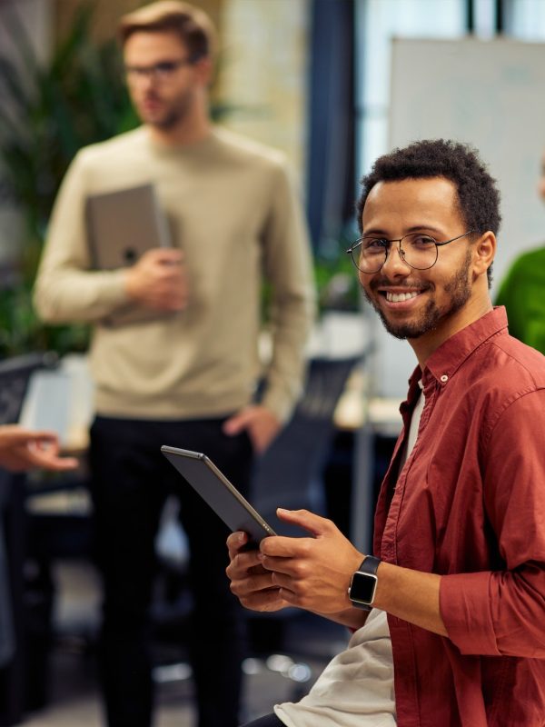 young-cheerful-mixed-race-man-holding-digital-tablet-and-smiling-at-camera-while-working-with.jpg